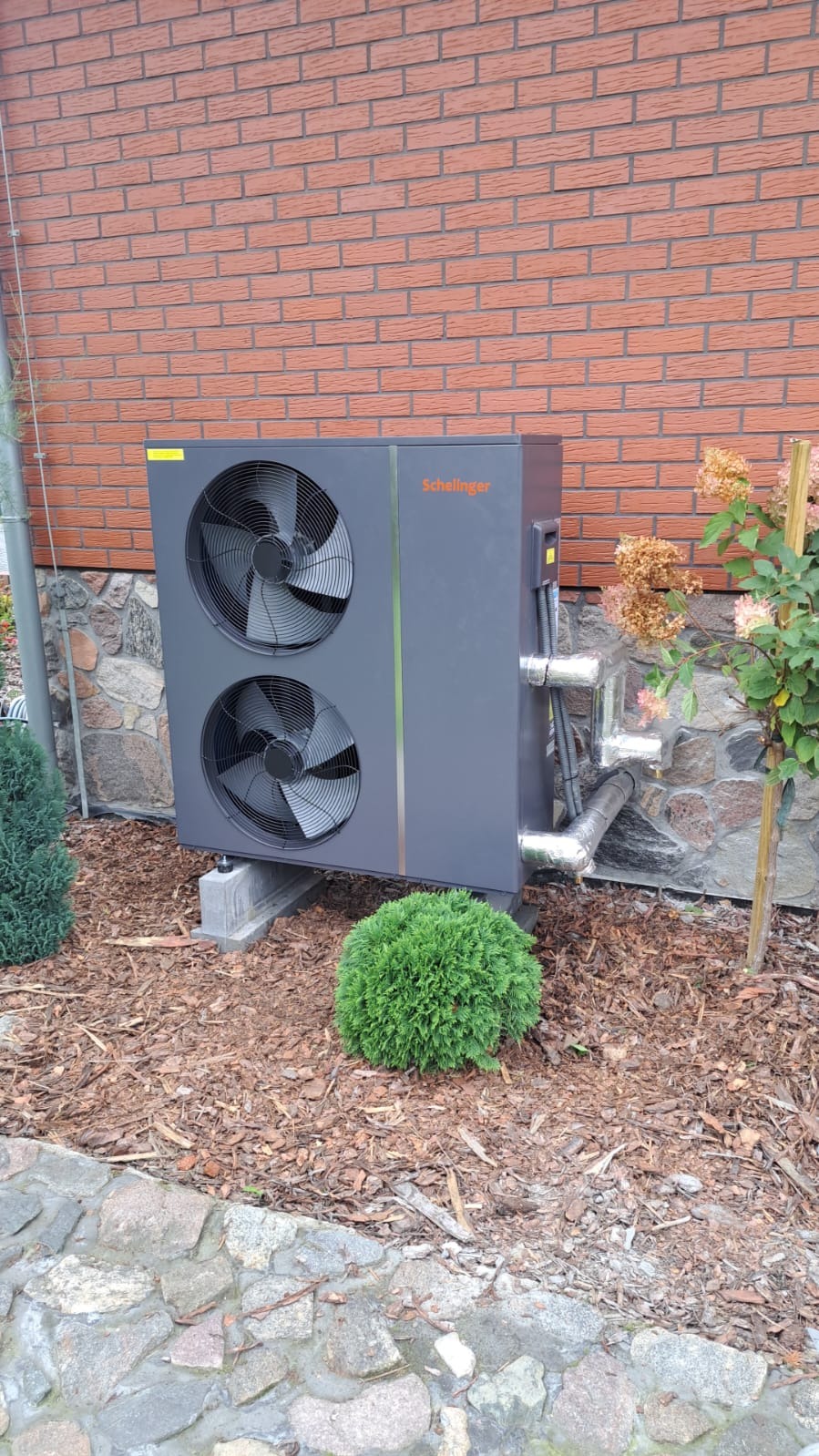 Zewnętrzna jednostka dwuwentylatorowa w kolorze grafitowym, zamontowana na betonowych podstawach przy ceglanym budynku z elementami kamiennymi, otoczona korą i niskimi krzewami.
