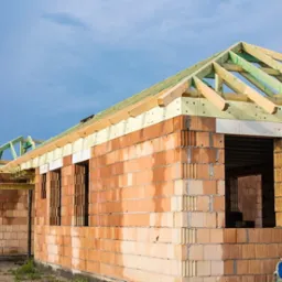 Budynek jednorodzinny w trakcie budowy: ściany z czerwonej cegły ceramicznej, drewniana konstrukcja dachu z zielonymi elementami, widoczne otwory okienne i drzwiowe, częściowo ocieplone ściany...