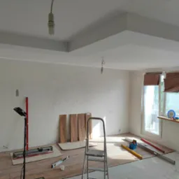 Wnętrze remontowanego pokoju z ułożoną częściowo podłogą z płytek imitujących drewno, widoczne narzędzia glazurnicze, drabina, lampa budowlana, oraz płyty podłogowe oparte o ścianę.