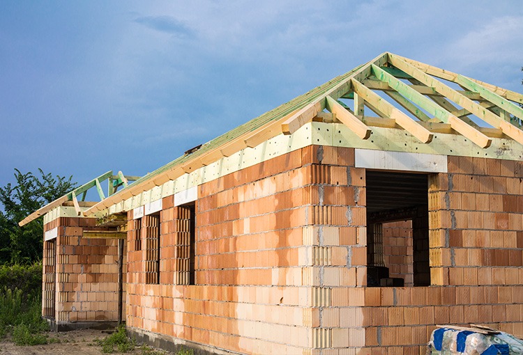 Budynek jednorodzinny w trakcie budowy: ściany z czerwonej cegły ceramicznej, drewniana konstrukcja dachu z zielonymi elementami, widoczne otwory okienne i drzwiowe, częściowo ocieplone ściany...