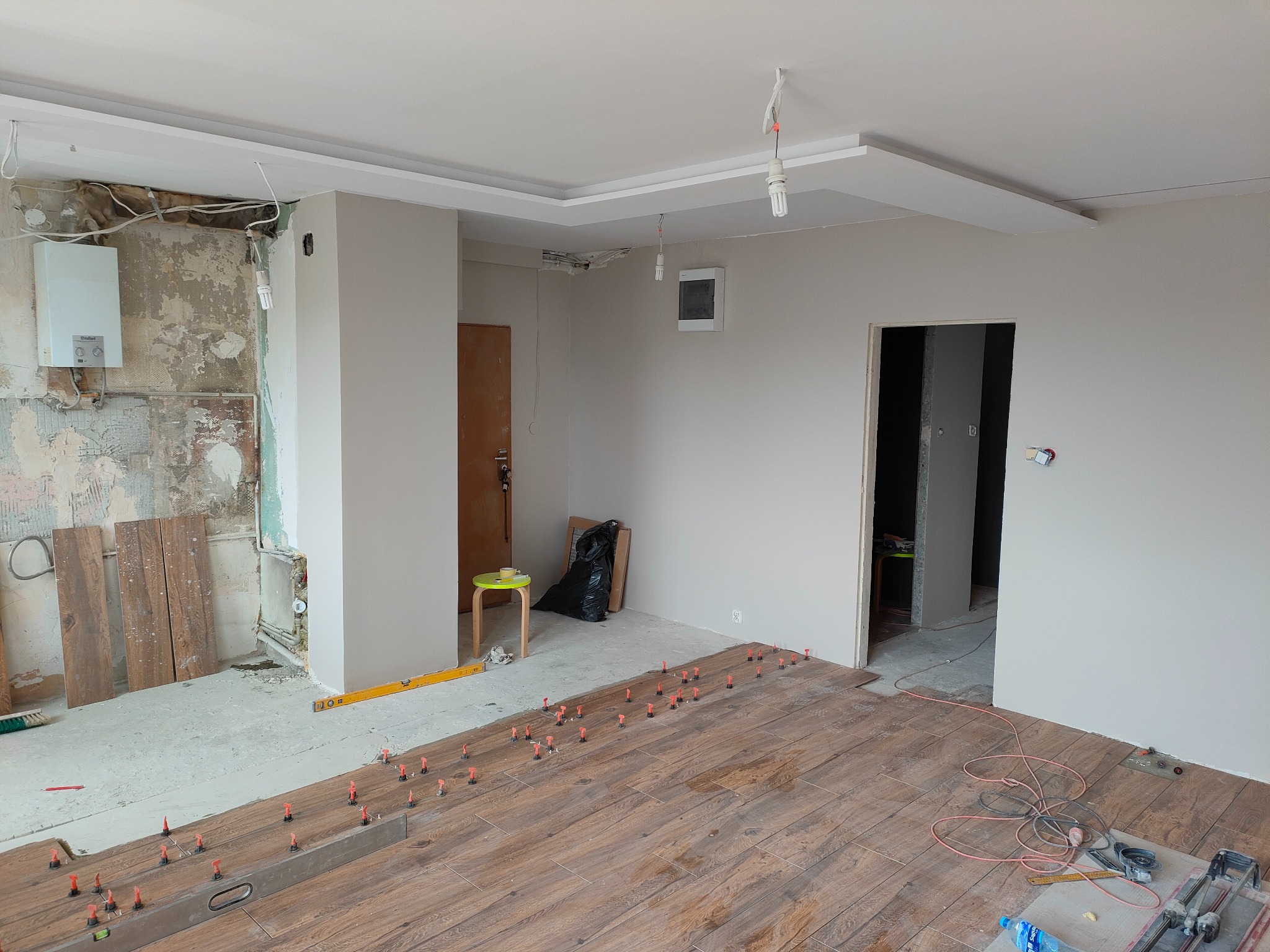 Wnętrze w trakcie remontu: układanie drewnopodobnych płytek podłogowych z użyciem systemu poziomowania, ściany częściowo otynkowane, widoczne instalacje, w tle krzesło i worek na śmieci.