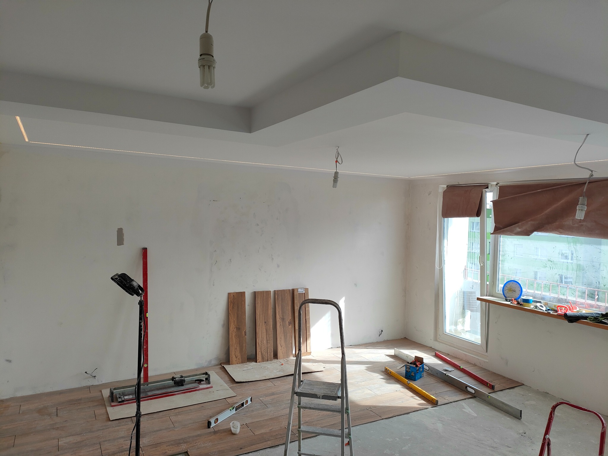 Wnętrze remontowanego pokoju z ułożoną częściowo podłogą z płytek imitujących drewno, widoczne narzędzia glazurnicze, drabina, lampa budowlana, oraz płyty podłogowe oparte o ścianę.