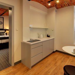 ARKADIUSZ DĘBICKI FIRMA BUDOWLANA - Nowoczesny aneks kuchenny z minimalistycznymi szarymi szafkami, białym blatem, płytą indukcyjną, okrągłym stołem z tulipanami w wazonie i widokiem na sypialnię z ceglanym sklepieniem.