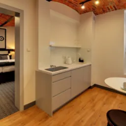 Nowoczesny aneks kuchenny z minimalistycznymi szarymi szafkami, białym blatem, płytą indukcyjną, okrągłym stołem z tulipanami w wazonie i widokiem na sypialnię z ceglanym sklepieniem.