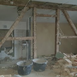 ARKADIUSZ DĘBICKI FIRMA BUDOWLANA - Wnętrze w trakcie demontażu ścianki działowej, widoczne dwa wiadra budowlane, gruz, odsłonięte instalacje elektryczne i fragment łazienki z umywalką.
