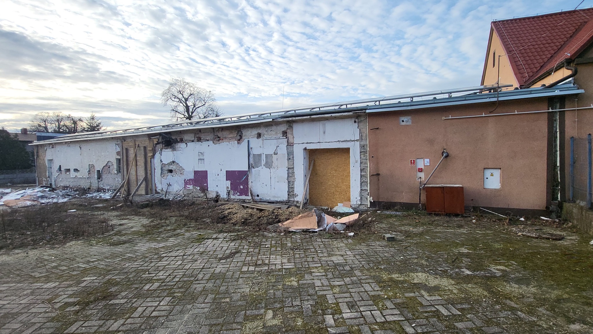 Budynek w trakcie remontu, z częściowo usuniętym tynkiem, odsłoniętą cegłą i zabezpieczonym otworem drzwiowym płytą OSB, na pierwszym planie gruz i błoto.