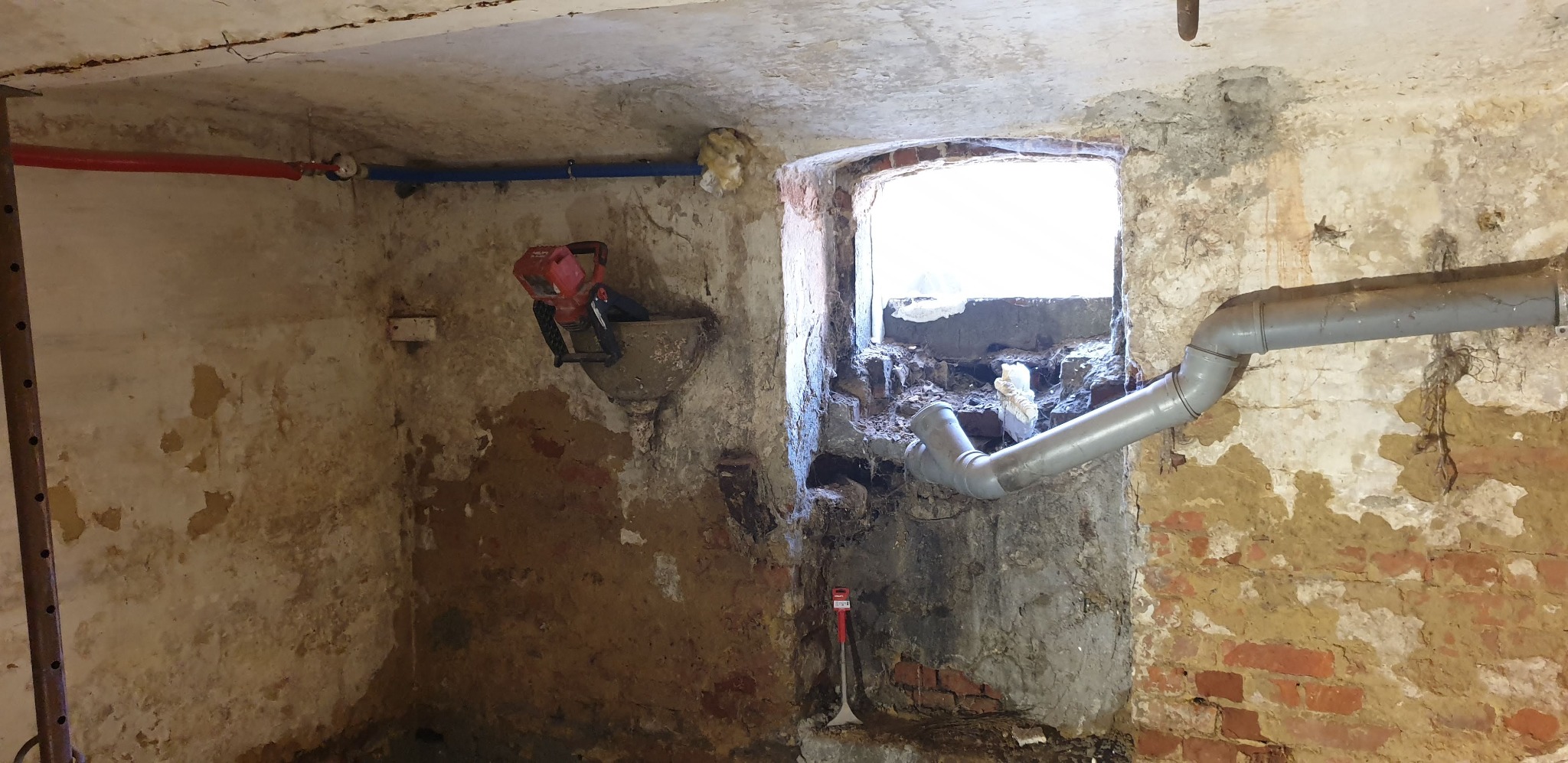 Piwnica w trakcie remontu: widoczne rury, uszkodzone ściany z fragmentami cegieł, okno z gruzem, wisząca mieszarka do zaprawy i szpachelka na podłodze.