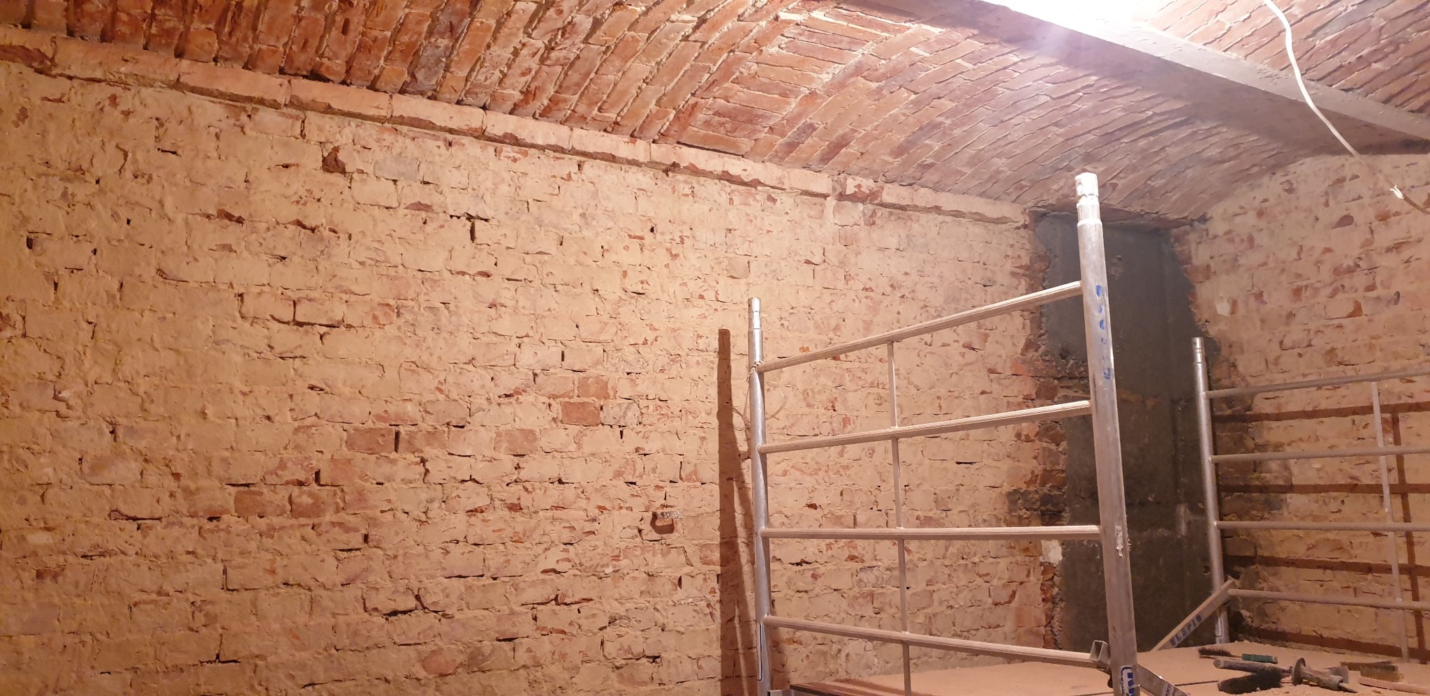 Ceglany sufit łukowy i ściana w trakcie remontu, częściowo pokryta szpachlą, z widocznym rusztowaniem i narzędziami na podłodze.