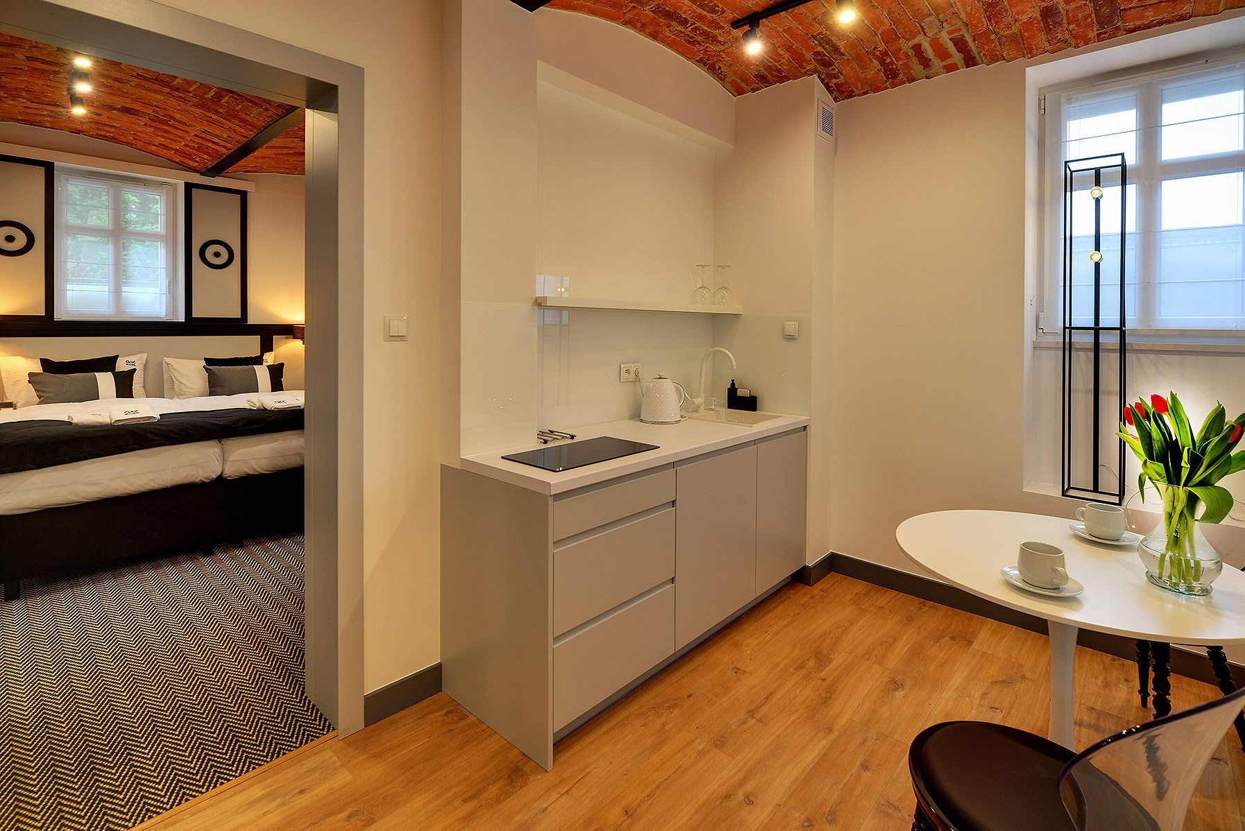 Nowoczesny aneks kuchenny z minimalistycznymi szarymi szafkami, białym blatem, płytą indukcyjną, okrągłym stołem z tulipanami w wazonie i widokiem na sypialnię z ceglanym sklepieniem.