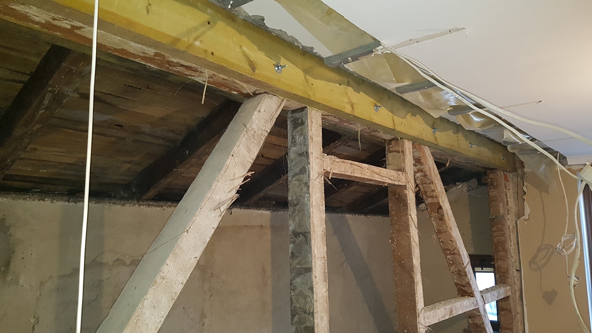 Wnętrze w trakcie remontu z odsłoniętą konstrukcją drewnianą stropu, widoczne elementy starej instalacji i przygotowania do montażu płyty kartonowo-gipsowej.