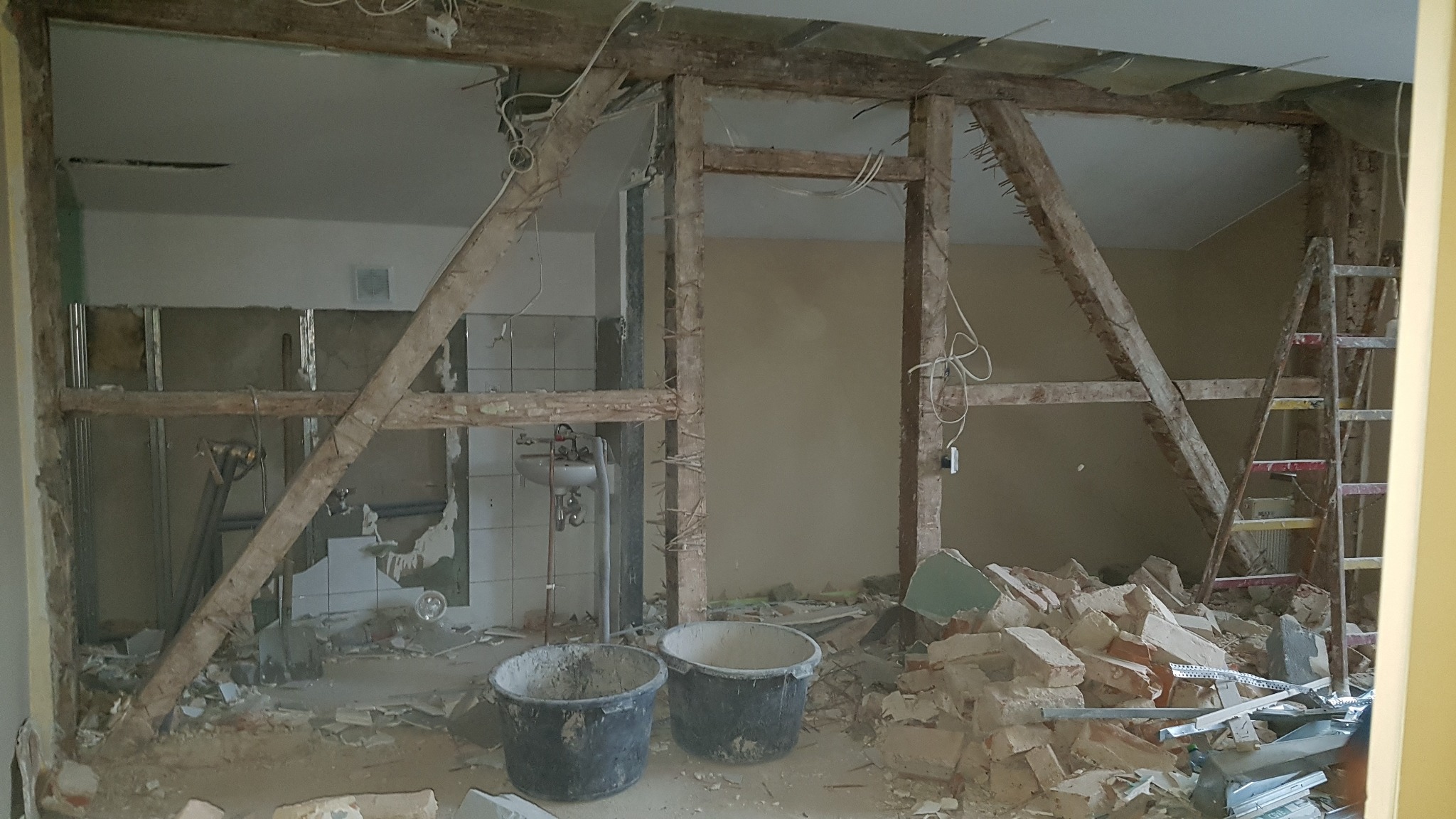 Wnętrze w trakcie demontażu ścianki działowej, widoczne dwa wiadra budowlane, gruz, odsłonięte instalacje elektryczne i fragment łazienki z umywalką.