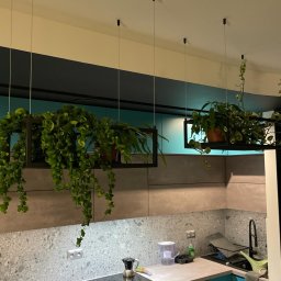 DTM - Nowoczesna kuchnia z turkusowymi szafkami, szarym blatem i kamienną ścianą, ozdobiona wiszącymi roślinami w czarnych, geometrycznych donicach podwieszonych na cienkich linkach.