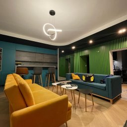 DTM - Nowoczesne wnętrze z otwartą kuchnią, gdzie turkusowe szafki kuchenne kontrastują z żółtą sofą i zieloną ścianą z pionowych paneli, podkreślone przez designerską lampę wiszącą.