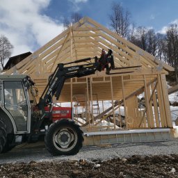 DTM - Montaż drewnianej konstrukcji domu za pomocą traktora z podnośnikiem teleskopowym na tle zimowego krajobrazu.