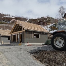 DTM - Wznoszenie domu w technologii szkieletowej: widok na konstrukcję z drewnianym szkieletem dachu i szarą elewacją, na tle górzystego krajobrazu z traktorem w prawym rogu.