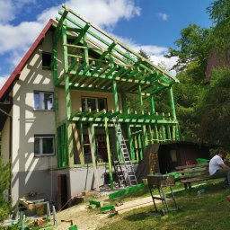 DTM - Wznoszenie dwukondygnacyjnego domu w konstrukcji szkieletowej z widocznymi, pomalowanymi na zielono elementami drewnianymi, drabiny opartymi o budynek, oraz osobą siedzącą na ławce obok grilla.