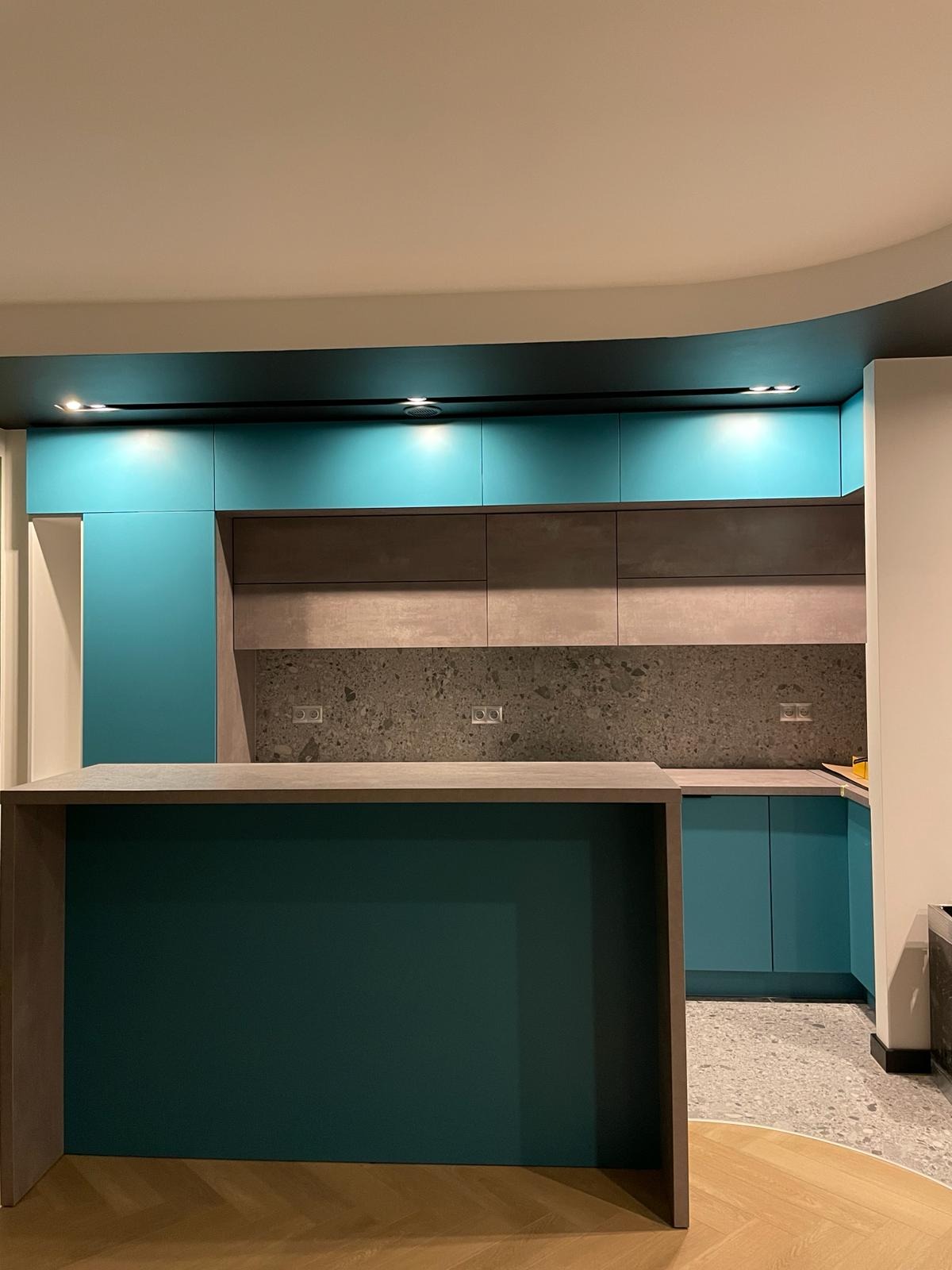 Nowoczesna kuchnia z wyspą, turkusowe i szare szafki, blat z szarego kamienia, parkiet w jodełkę.