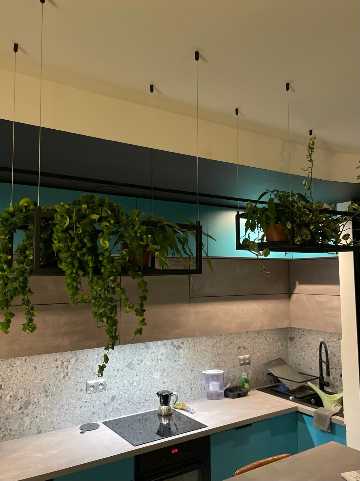 Nowoczesna kuchnia z turkusowymi szafkami, szarym blatem i kamienną ścianą, ozdobiona wiszącymi roślinami w czarnych, geometrycznych donicach podwieszonych na cienkich linkach.