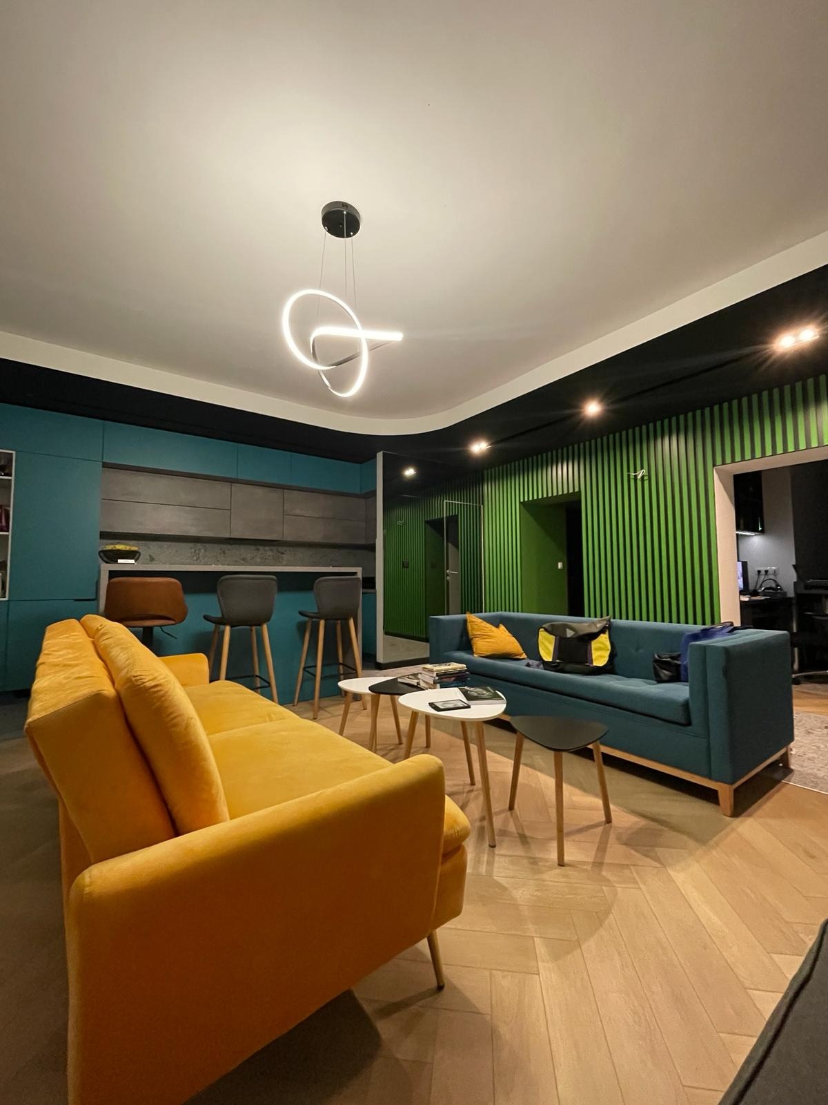 Nowoczesne wnętrze z otwartą kuchnią, gdzie turkusowe szafki kuchenne kontrastują z żółtą sofą i zieloną ścianą z pionowych paneli, podkreślone przez designerską lampę wiszącą.