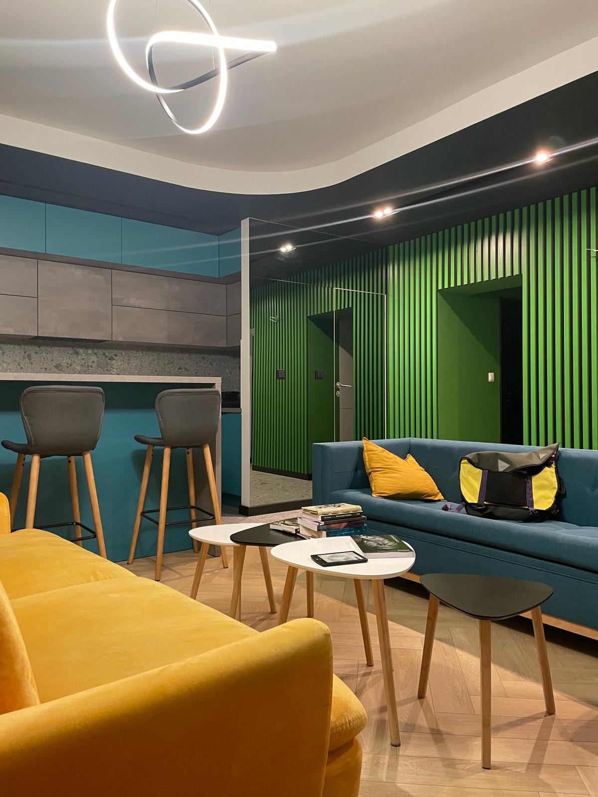 Nowoczesne wnętrze z żółtą sofą, turkusową kuchnią z hokerami, zieloną ścianą z pionowych paneli i okrągłą lampą sufitową.