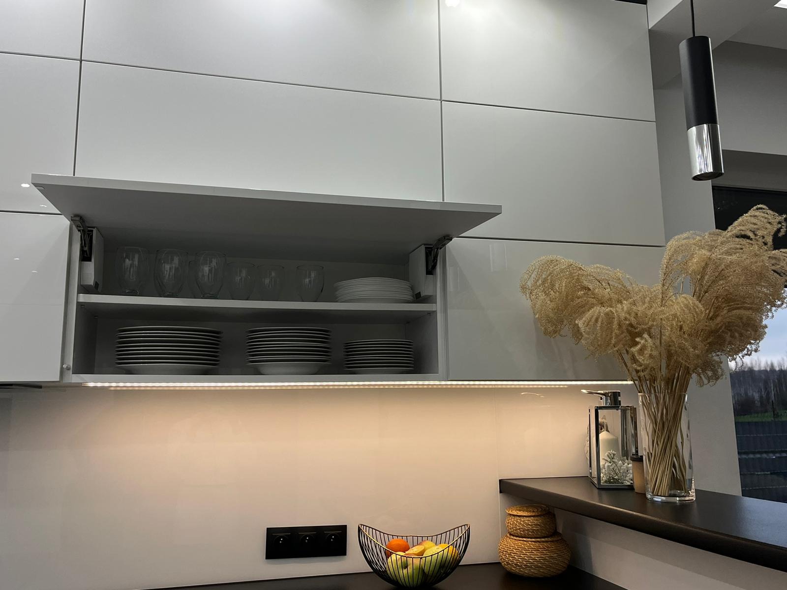 Minimalistyczna kuchnia: białe, lakierowane szafki z otwartą szafką górną, podświetlenie LED, dekoracje z suszonych traw w wazonie i miska z owocami na blacie.