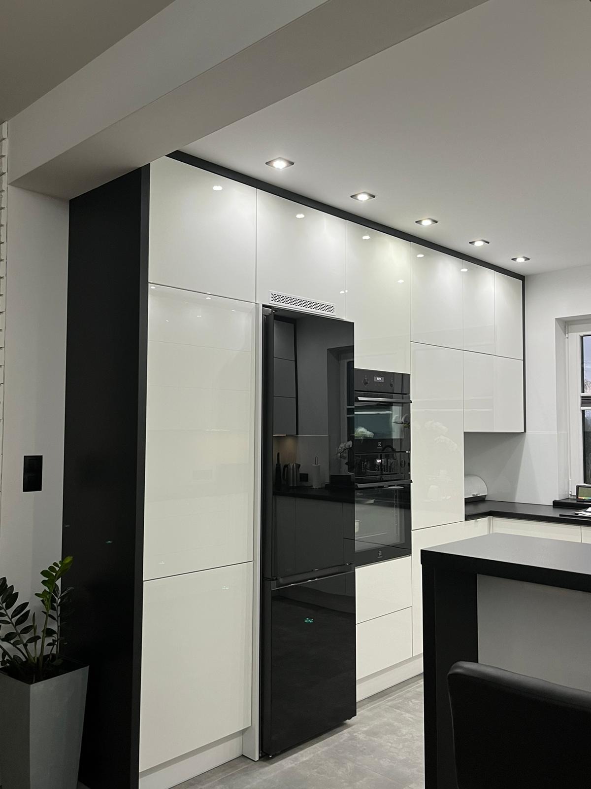 Nowoczesna kuchnia z białymi, lakierowanymi frontami szafek i czarnymi akcentami, wbudowana lodówka z czarnym frontem, oświetlenie punktowe podszafkowe i sufitowe.