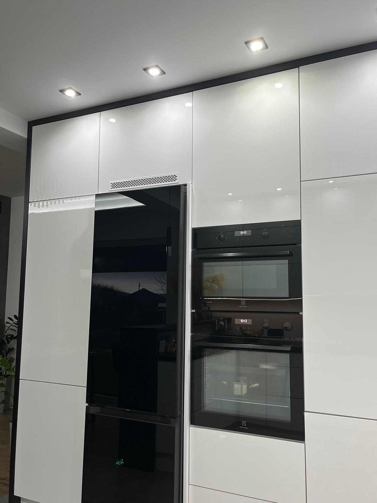 Nowoczesna zabudowa kuchenna z białymi, lakierowanymi frontami, czarnymi obramowaniami, wbudowaną czarną lodówką i piekarnikiem z mikrofalówką.