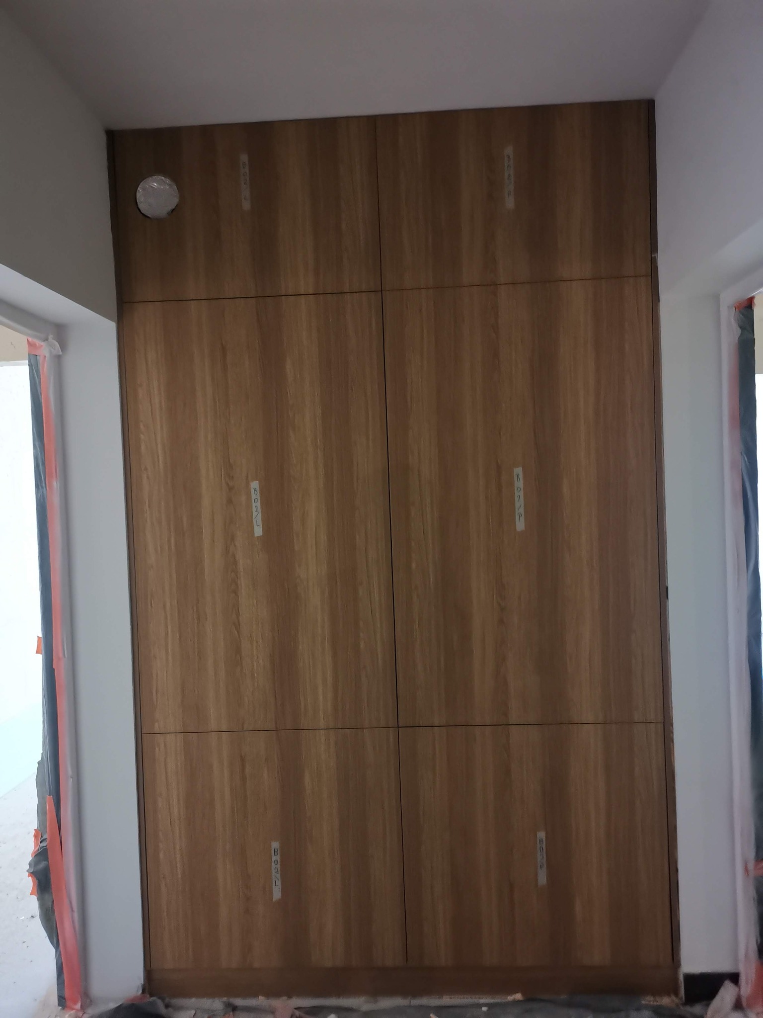 Wysoka, drewniana szafa wnękowa z sześcioma drzwiami o usłojeniu w odcieniach brązu, widoczna podczas montażu, z taśmami zabezpieczającymi na krawędziach i otworem na instalację elektryczną...
