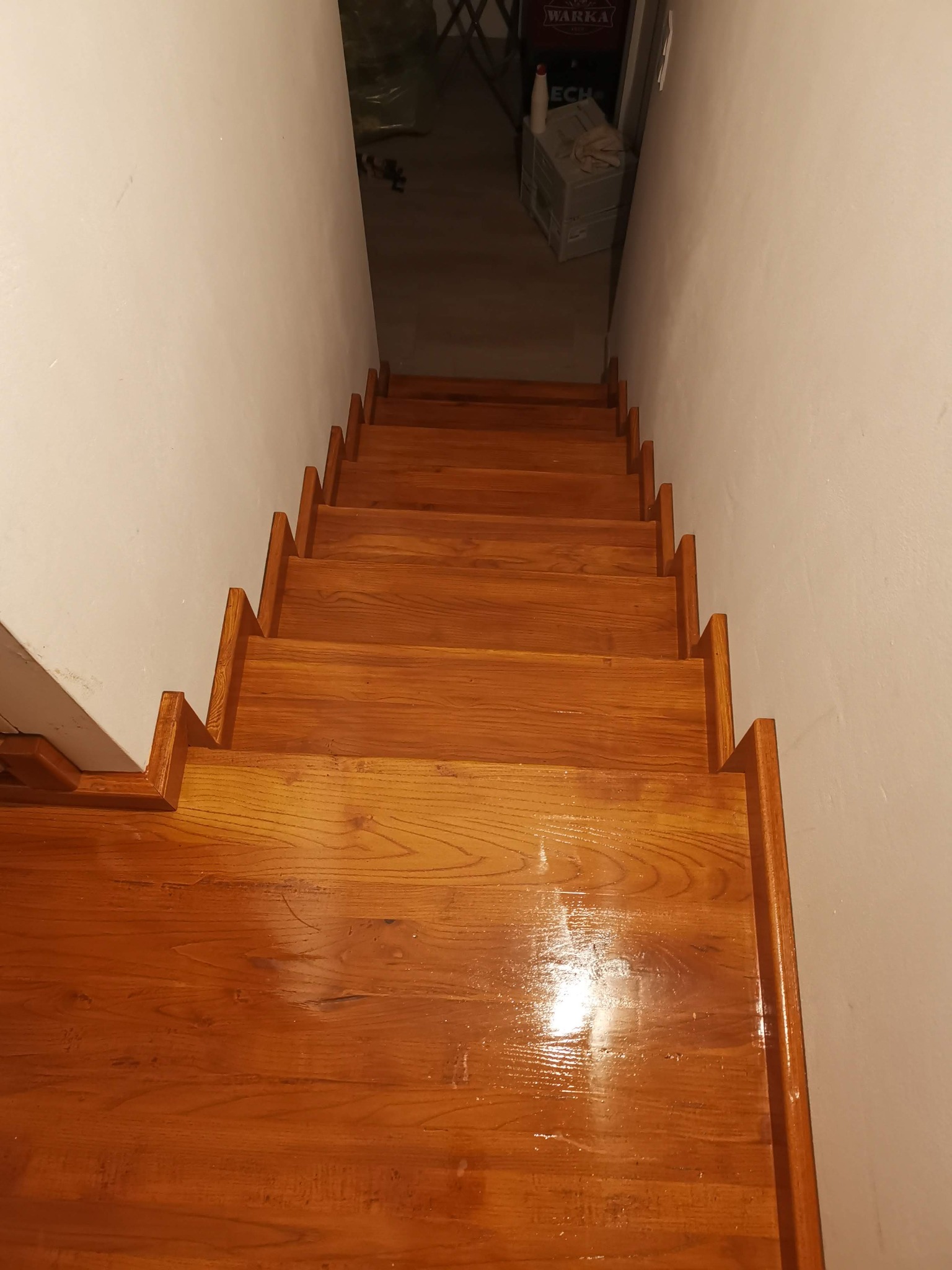 Drewniane schody w dół, lakierowane na wysoki połysk, widok z góry, ściany pomalowane na biało, w tle widoczna skrzynka z napojami.