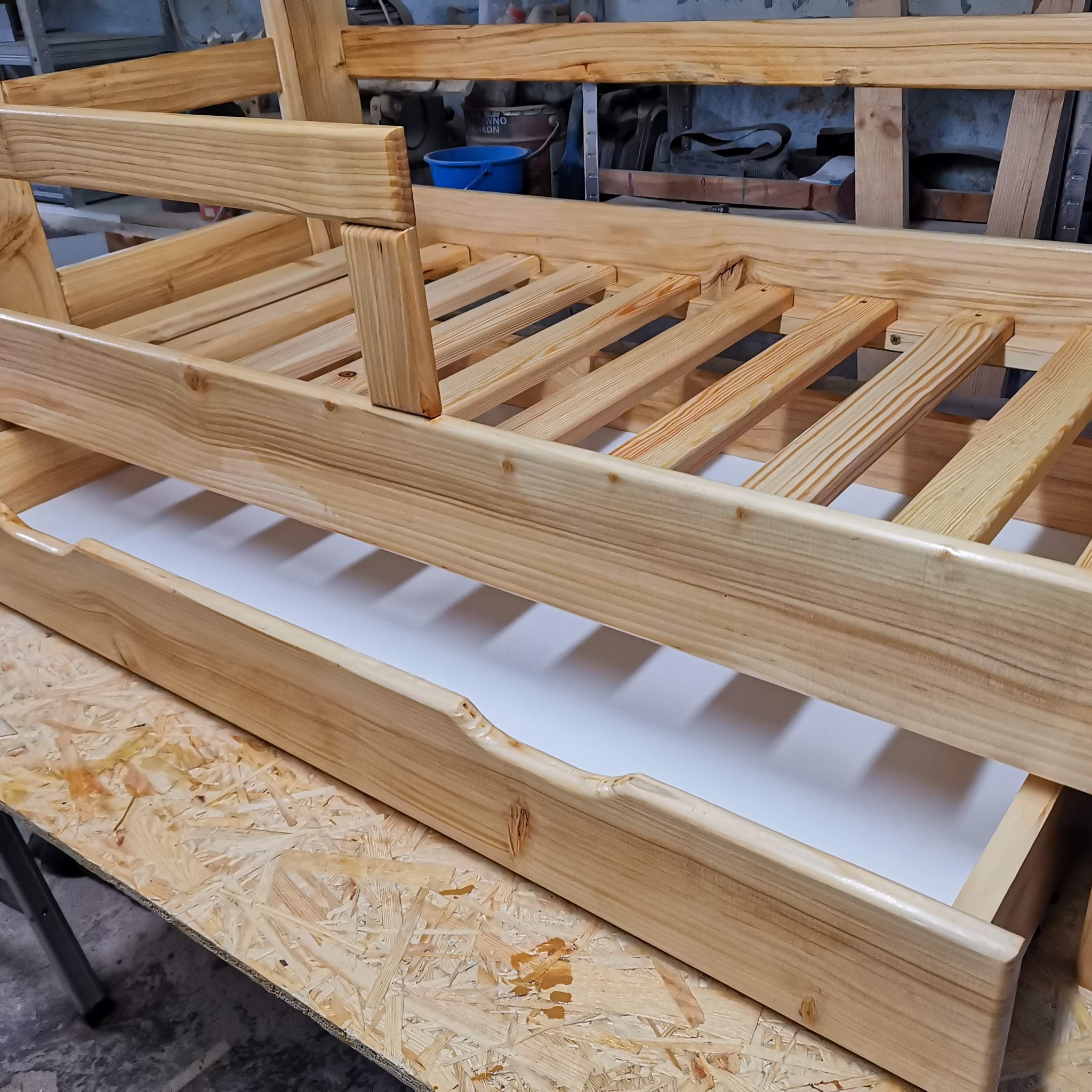 Drewniana konstrukcja łóżka, prawdopodobnie w trakcie montażu, z widocznymi elementami ramy i stelaża, surowe drewno z widocznymi słojami, na tle warsztatu.