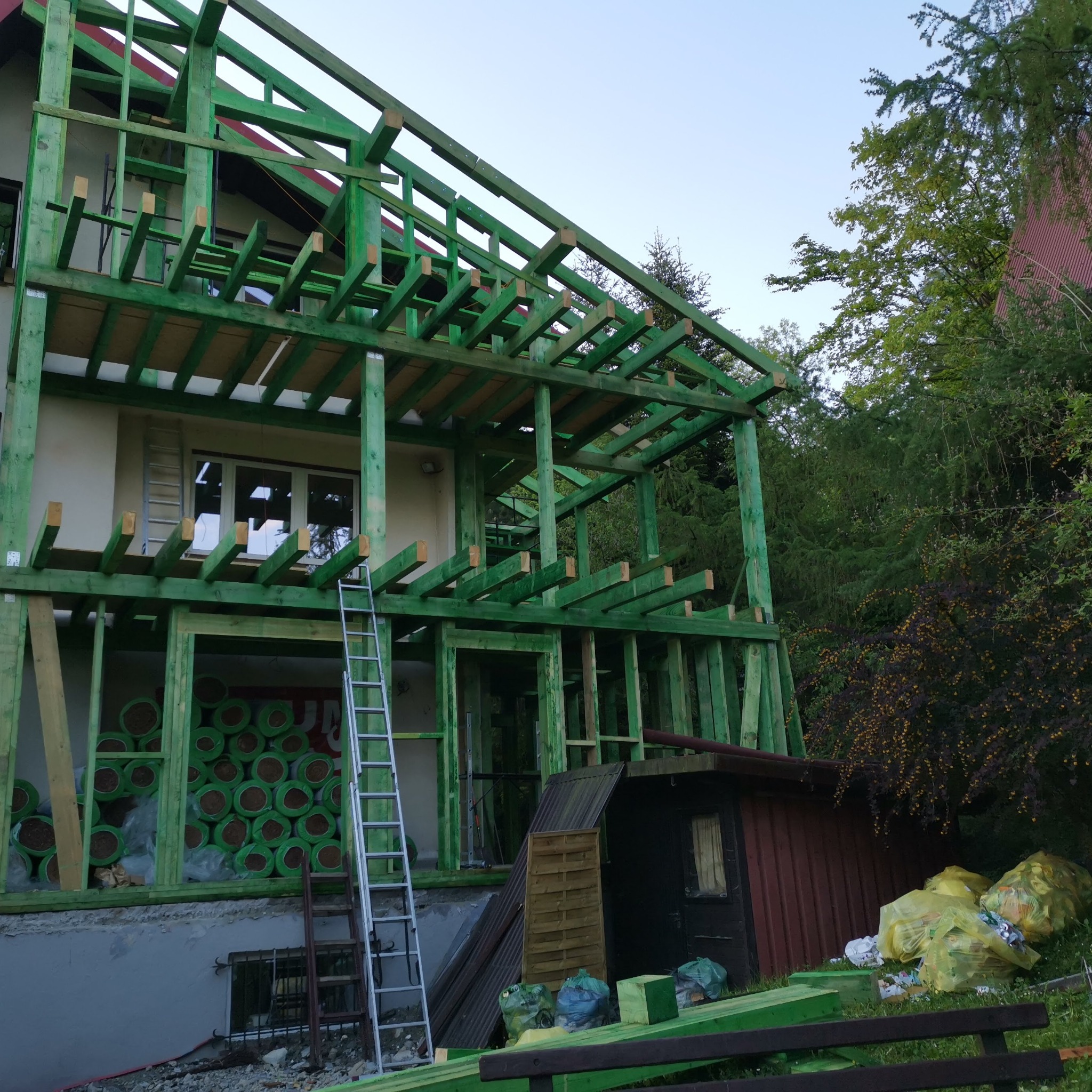 Surowa, zielona drewniana konstrukcja domu w trakcie budowy, widoczne rolki izolacji wewnątrz, drabina oparta o ścianę, otoczenie w stanie budowy.