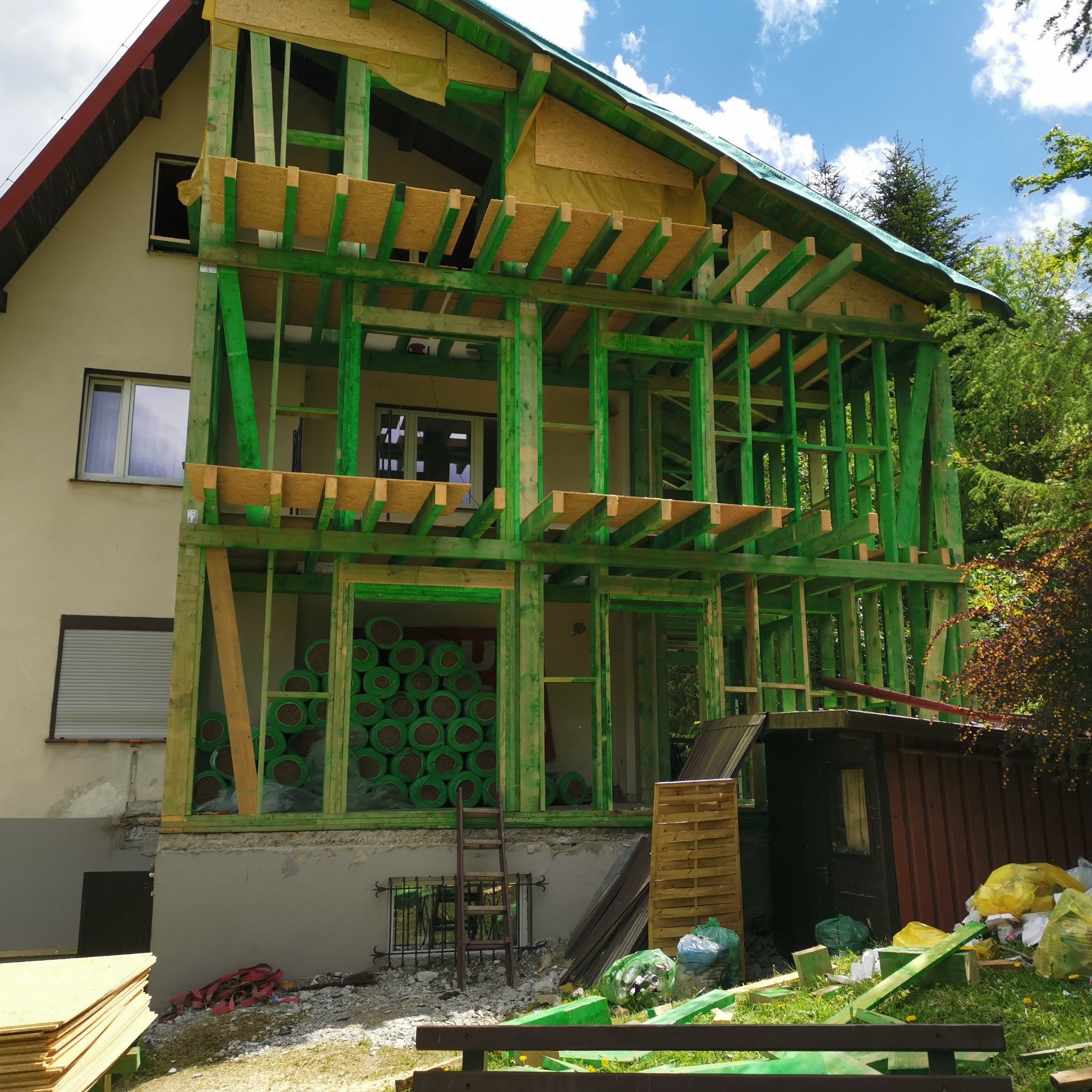 Budowa domu w technologii szkieletowej: widoczna konstrukcja drewniana pomalowana na zielono, częściowo obudowana płytami OSB, z rolkami izolacji wewnątrz. Budynek dwukondygnacyjny, w tle drzewa...