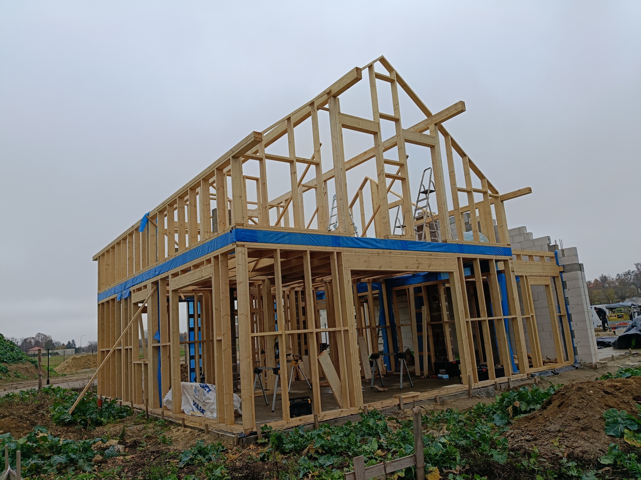 Surowy szkielet domu w trakcie budowy, z widocznymi drewnianymi belkami i niebieską folią ochronną. W tle pochmurne niebo i częściowo widoczne otoczenie budowy.