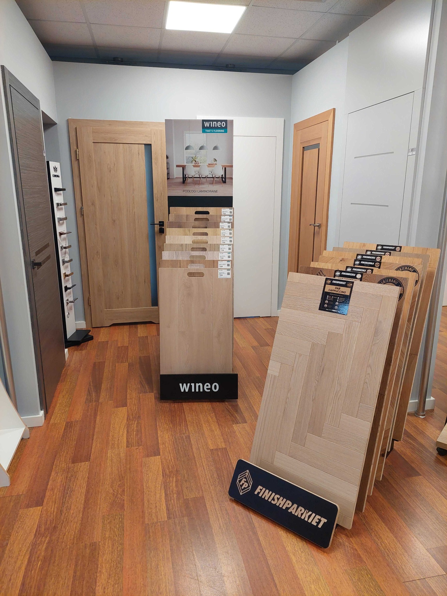 Ekspozycja w salonie z różnymi modelami drzwi i paneli podłogowych marek Wineo i Finishparkiet, z widocznym wzorem ułożenia w jodełkę na panelach.