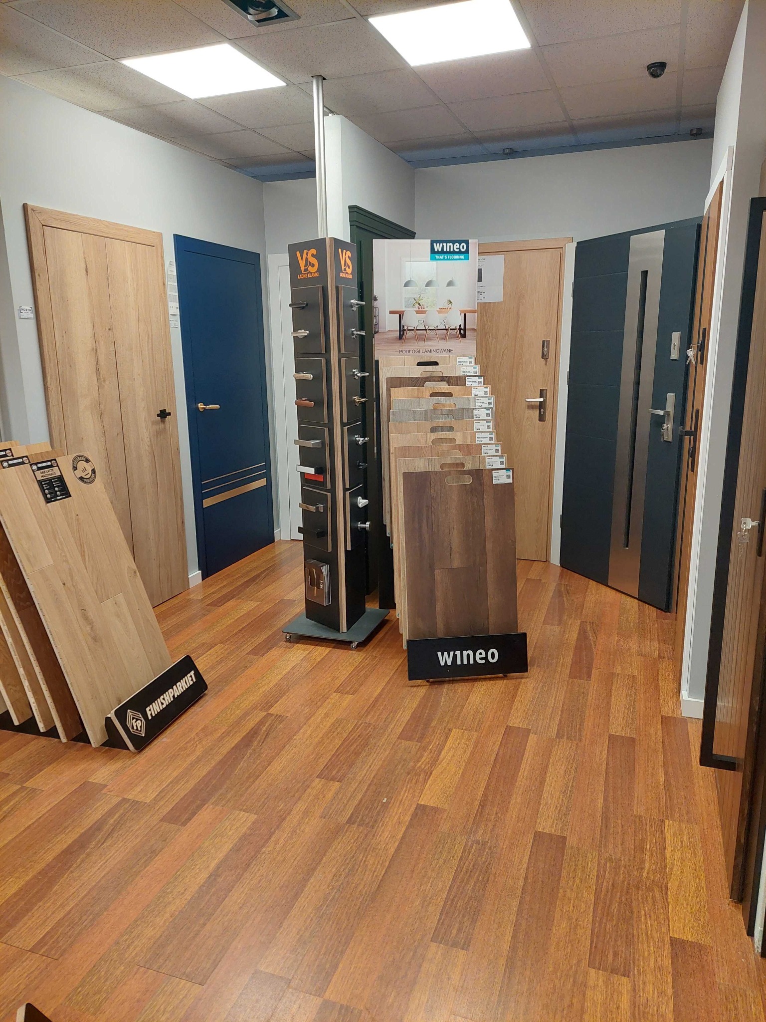 Ekspozycja w salonie z różnymi modelami drzwi i paneli podłogowych, widoczne drzwi w kolorze drewna, niebieskie oraz stalowe, stojak z klamkami i ekspozytory paneli laminowanych marek Wineo...