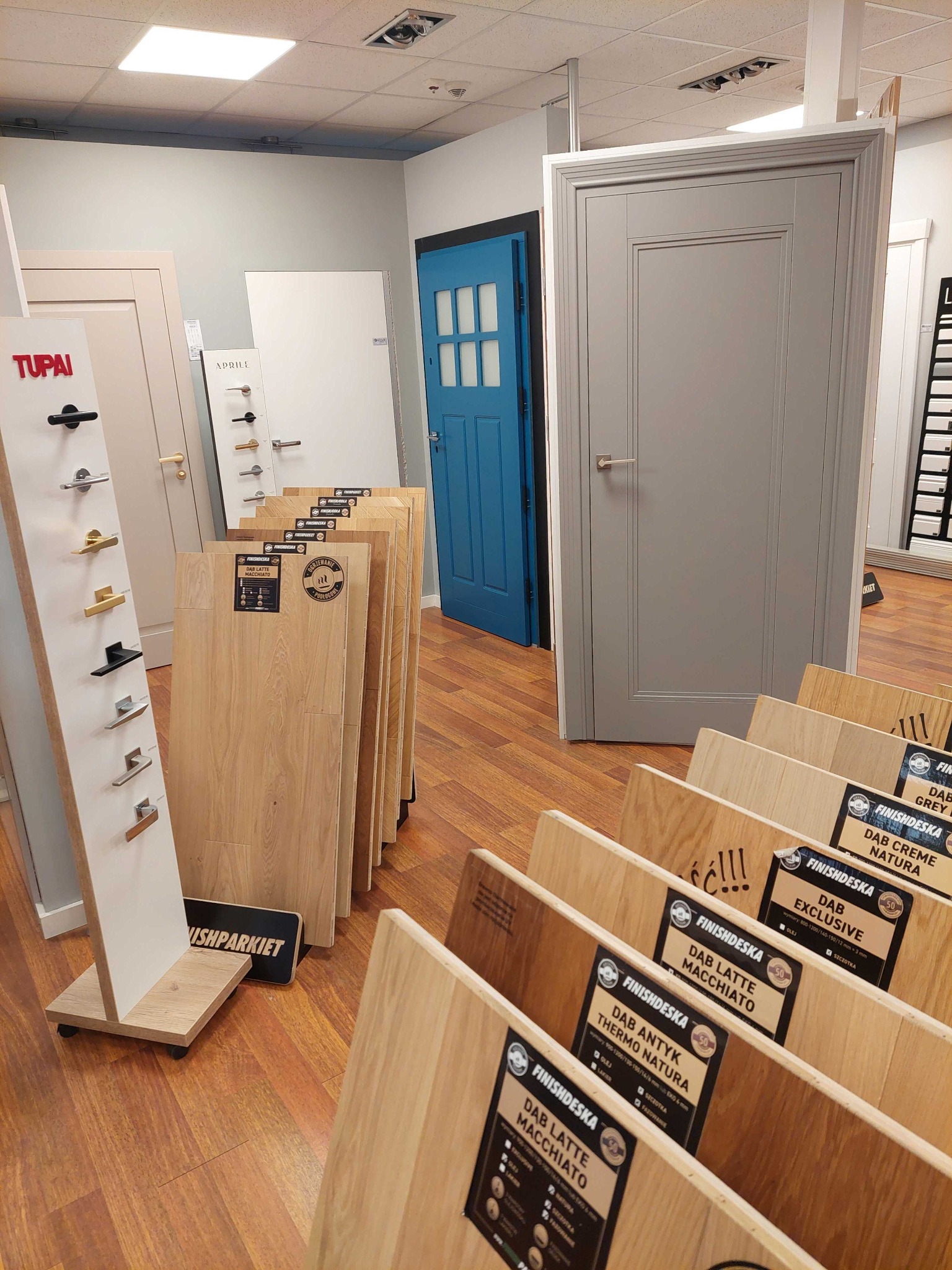 Ekspozycja drzwi wewnętrznych w salonie: szare drzwi z prostokątnymi panelami, obok niebieskie drzwi z szybkami, oraz panele podłogowe z opisami gatunków drewna i uchwyty do drzwi na stojaku.