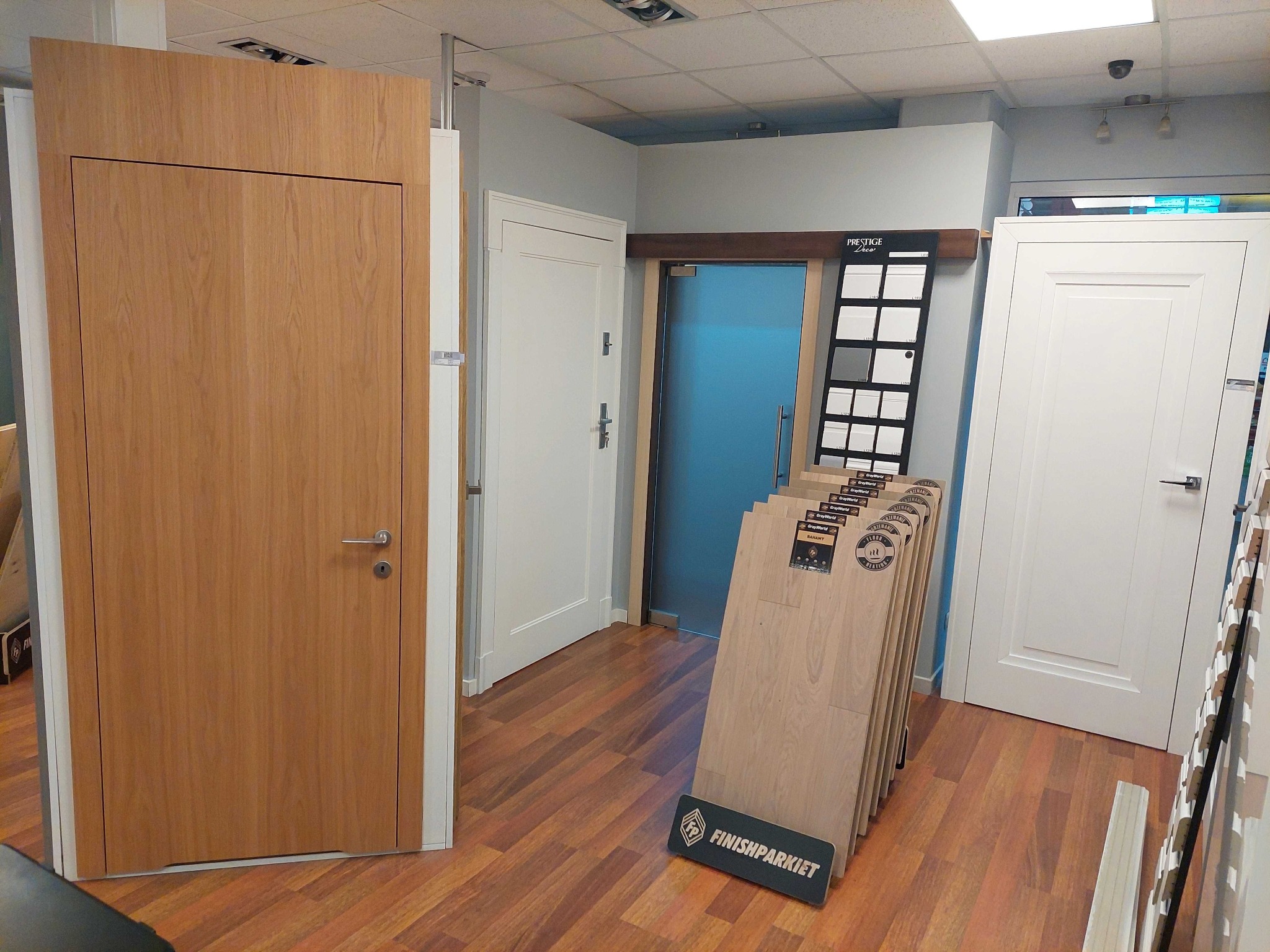 Ekspozycja w salonie: jasne skrzydło drzwiowe z widocznym usłojeniem, obok próbki paneli podłogowych i inne modele drzwi w tle.