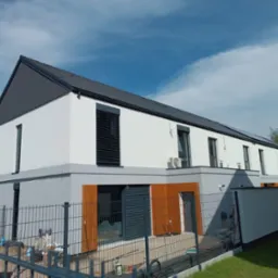 Nowoczesny budynek szeregowy z białą elewacją, szarym dachem i drewnianymi akcentami, widok z zewnątrz przez metalowe ogrodzenie.