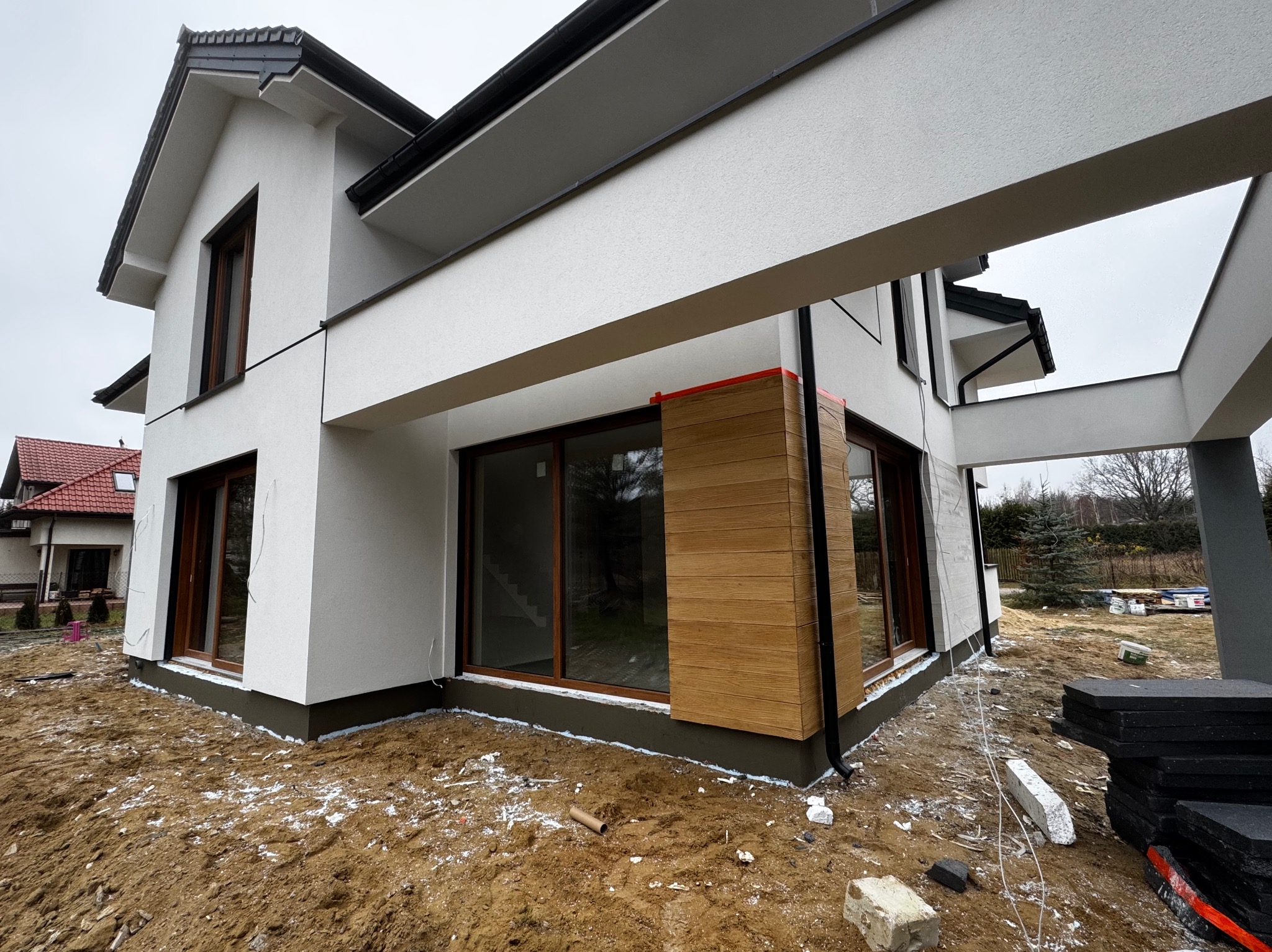 Nowoczesny dom jednorodzinny z białą elewacją i drewnianą okładziną, widoczne prace wykończeniowe wokół budynku, ocieplenie fundamentów i przygotowanie terenu.