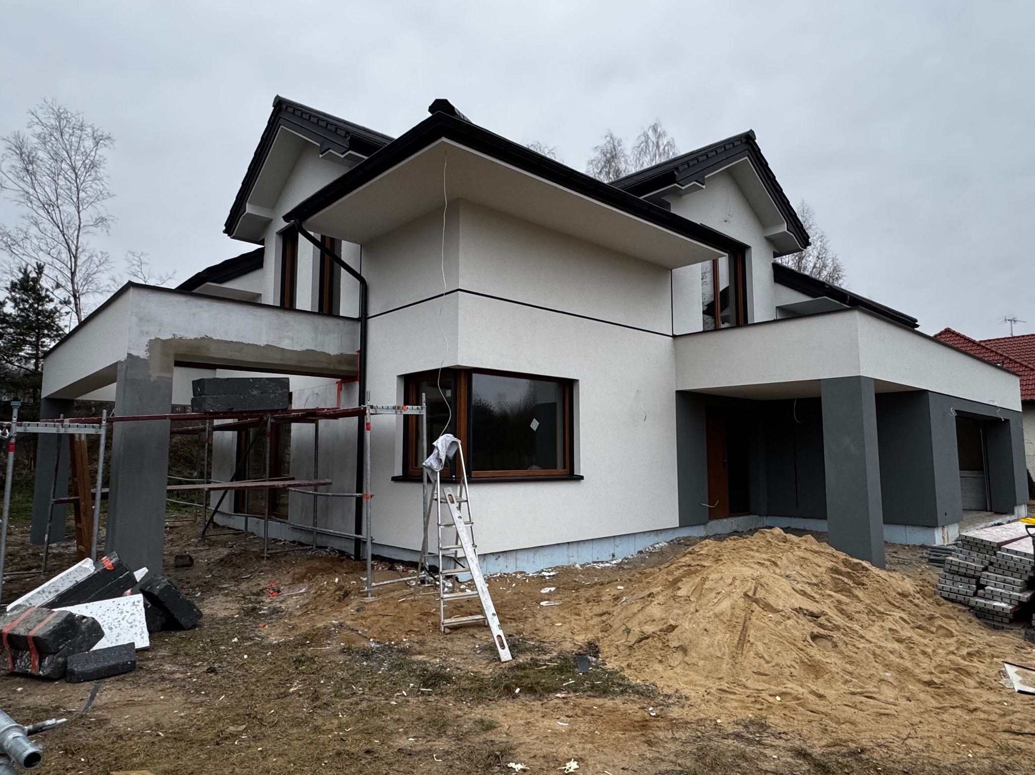 Dom jednorodzinny w Żyrardowie w trakcie prac elewacyjnych; widoczne rusztowania, stos materiałów budowlanych i hałda piasku. Część elewacji jest już wykończona w kolorze białym, a część w szarym.