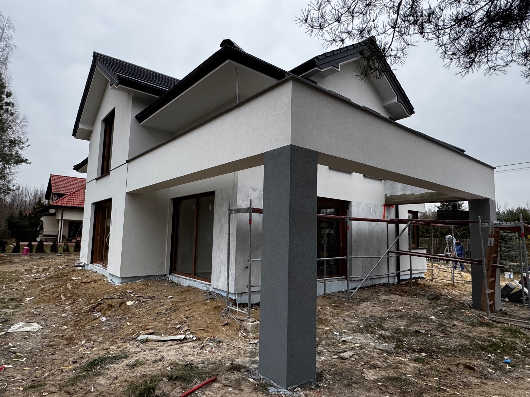 Nowoczesny dom jednorodzinny w trakcie prac wykończeniowych elewacji, widoczne rusztowania i pracownik w niebieskiej kurtce. Częściowo otynkowana fasada w kolorze białym, szara kolumna...