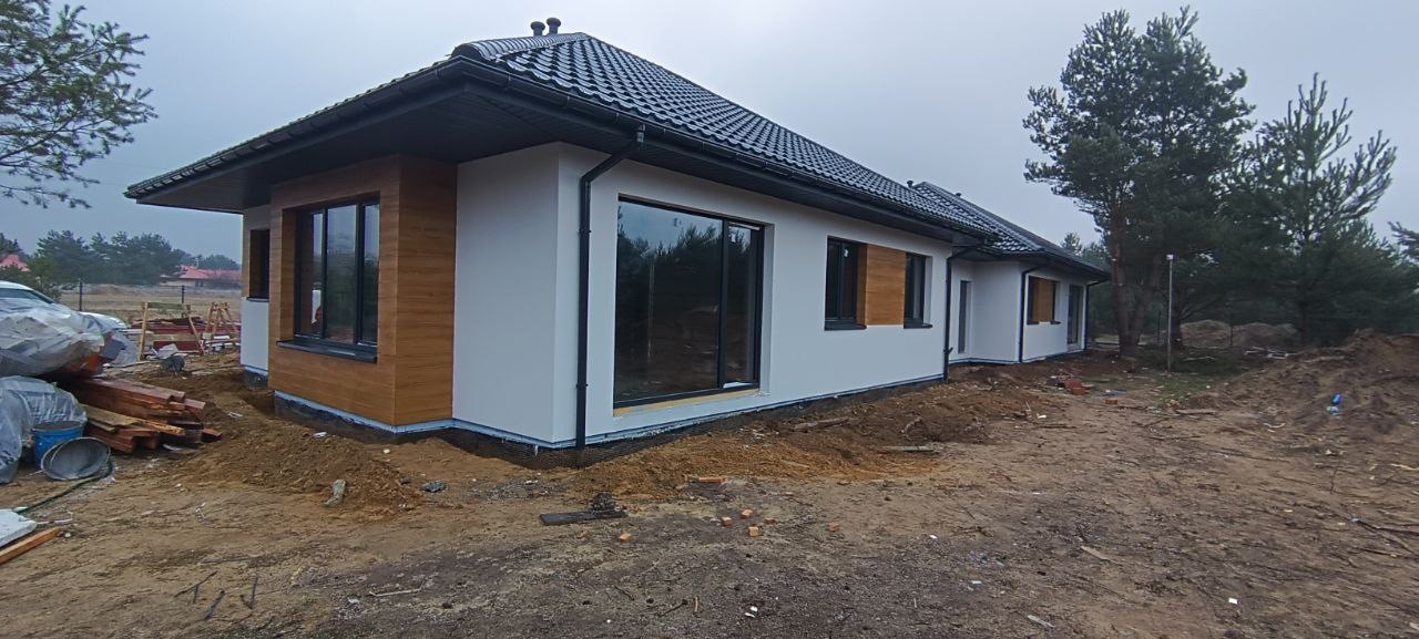 Nowoczesny parterowy dom z białą elewacją i drewnopodobnymi panelami elewacyjnymi, widoczne przygotowania terenu wokół budynku.