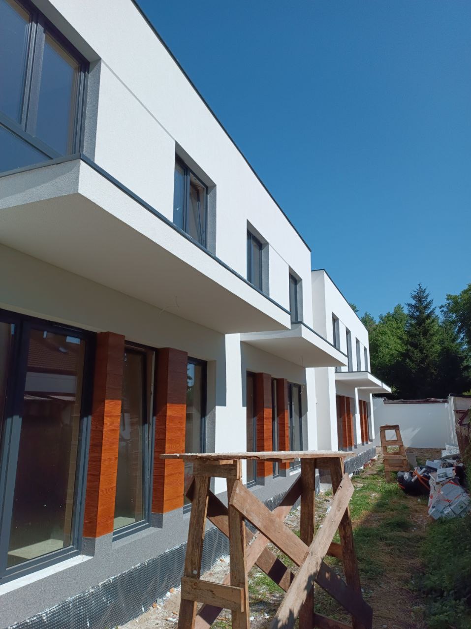 Nowoczesny dom w Żyrardowie z białą elewacją, drewnopodobnymi elementami dekoracyjnymi wokół okien i szarą podmurówką, widoczny fragment rusztowania.