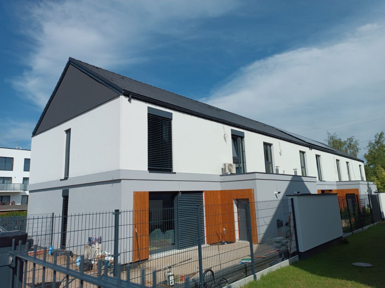 Nowoczesny budynek szeregowy z białą elewacją, szarym dachem i drewnianymi akcentami, widok z zewnątrz przez metalowe ogrodzenie.