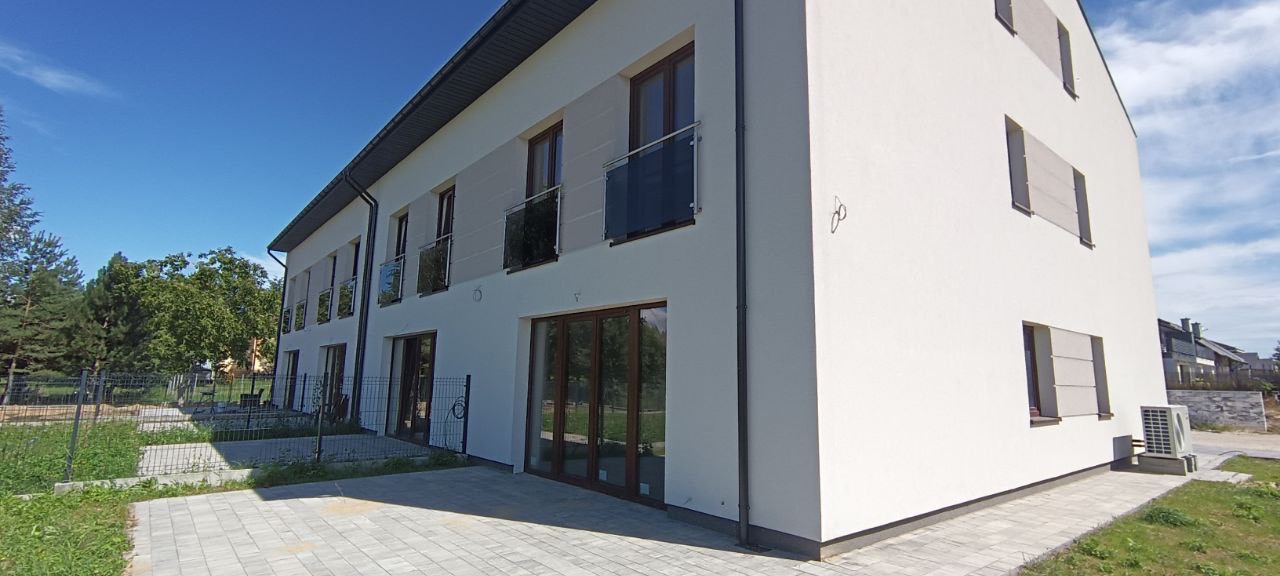 Nowoczesny budynek wielorodzinny z jasną, gładką elewacją, szklanymi balkonami i brązowymi ramami okiennymi, sfotografowany w słoneczny dzień.