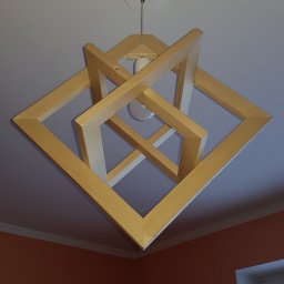 Nowoczesna lampa wisząca z jasnego drewna, składająca się z kwadratowych ram, zawieszona na białym suficie. Widoczna żarówka i fragment pomarańczowej ściany.