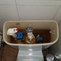 Wnętrze spłuczki toalety z widocznymi elementami mechanicznymi, woda o brązowym zabarwieniu, białe płytki w tle. Ujęcie z góry.