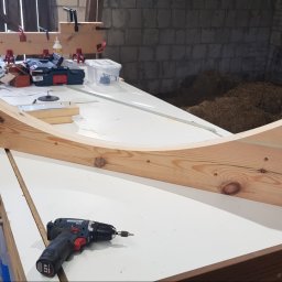 Element z jasnego drewna o zakrzywionym kształcie na stole warsztatowym, obok wiertarka. W tle narzędzia i bele siana. Detal stolarski w surowym warsztacie.