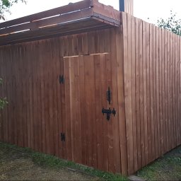 Adam Wasilewski - Drewniana konstrukcja z zadaszeniem, prawdopodobnie szopa lub mały budynek gospodarczy, wykonana z pionowo ułożonych desek, z czarnym zamkiem na drzwiach.