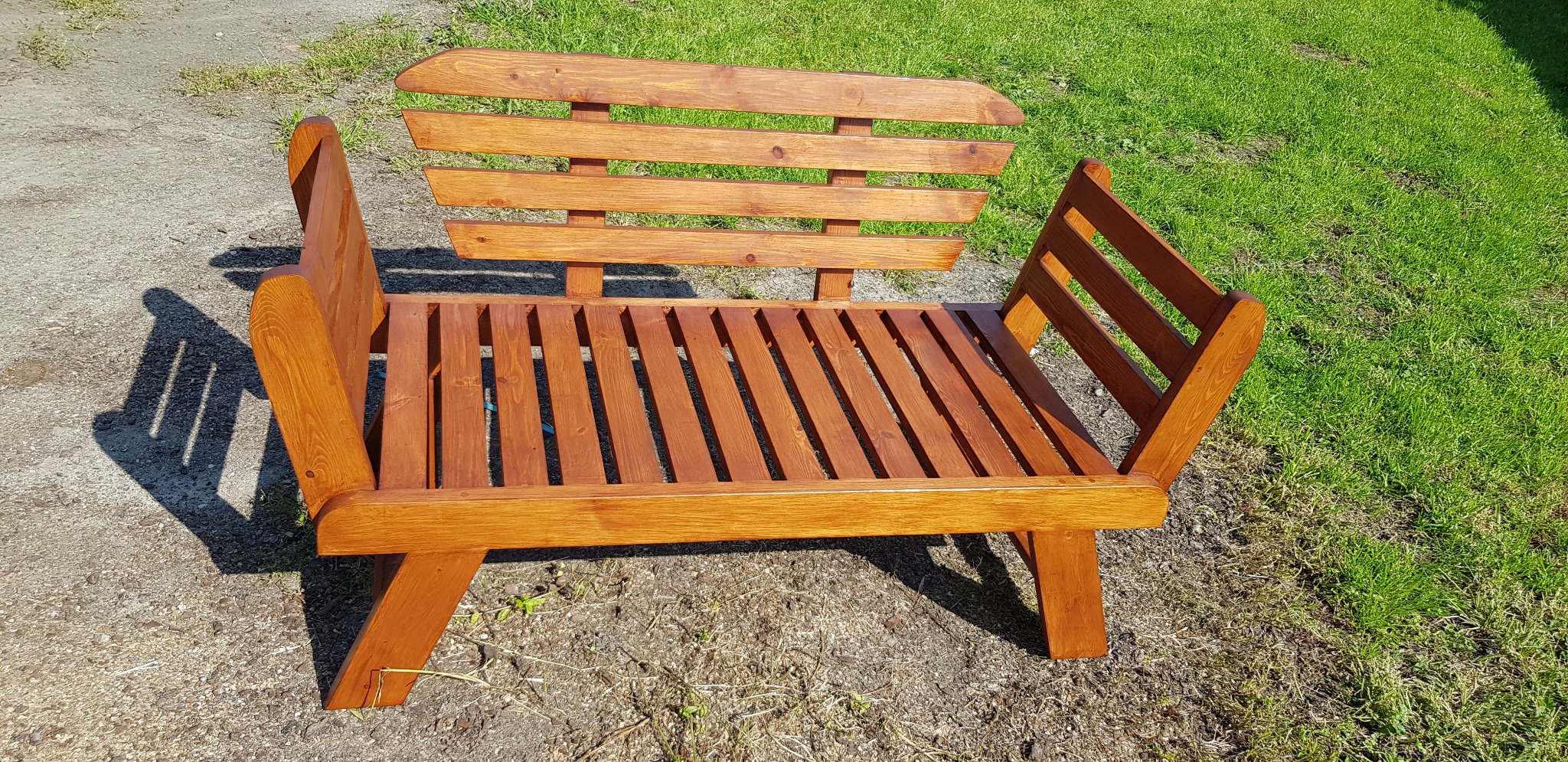 Drewniana ławka ogrodowa z oparciem i podłokietnikami, pomalowana na brązowo, stoi na trawie. Widoczne detale konstrukcyjne i naturalne usłojenie drewna.